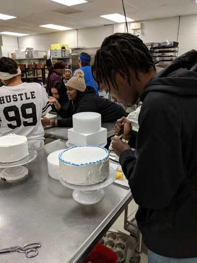Male student decorating a cake around the top with blue striped buttercream frosting.