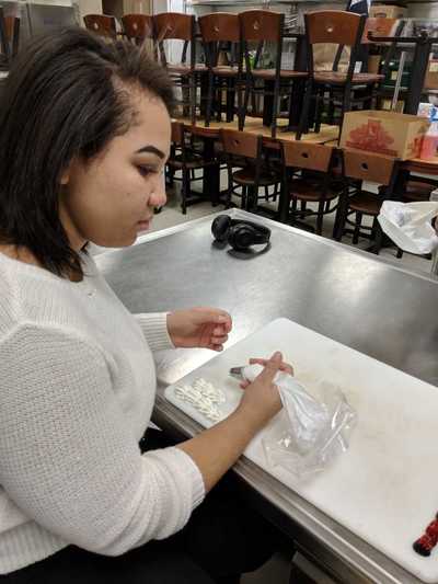 Female student using a pastry bag to squeeze out white buttercream frosting to practice decorating on a  cutting board.