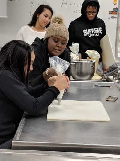 Two female students, one viewing and one using a pastry bag filled with white buttercream frosting to practice decorating techniques.
