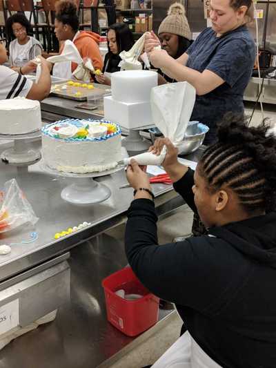 Female student decorating a cake by adding a border of white buttercream frosting at bottom.  