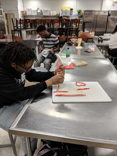 Male student using red buttercream frosting to practice cake decorating techniques.