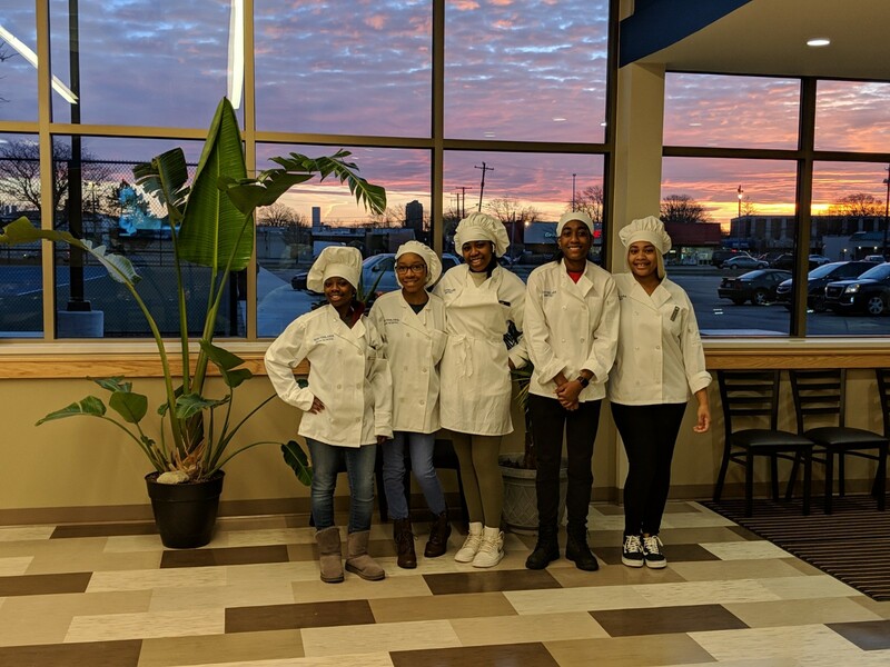 Student chefs posing in front of sunrise in bistro window.