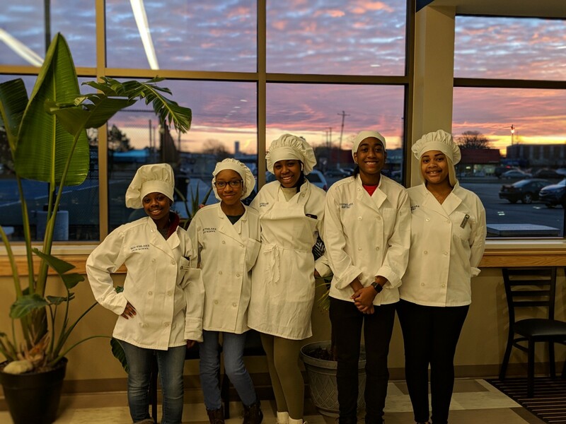 Five student chefs posing in front of a beautiful sunrise in the bistro window.  